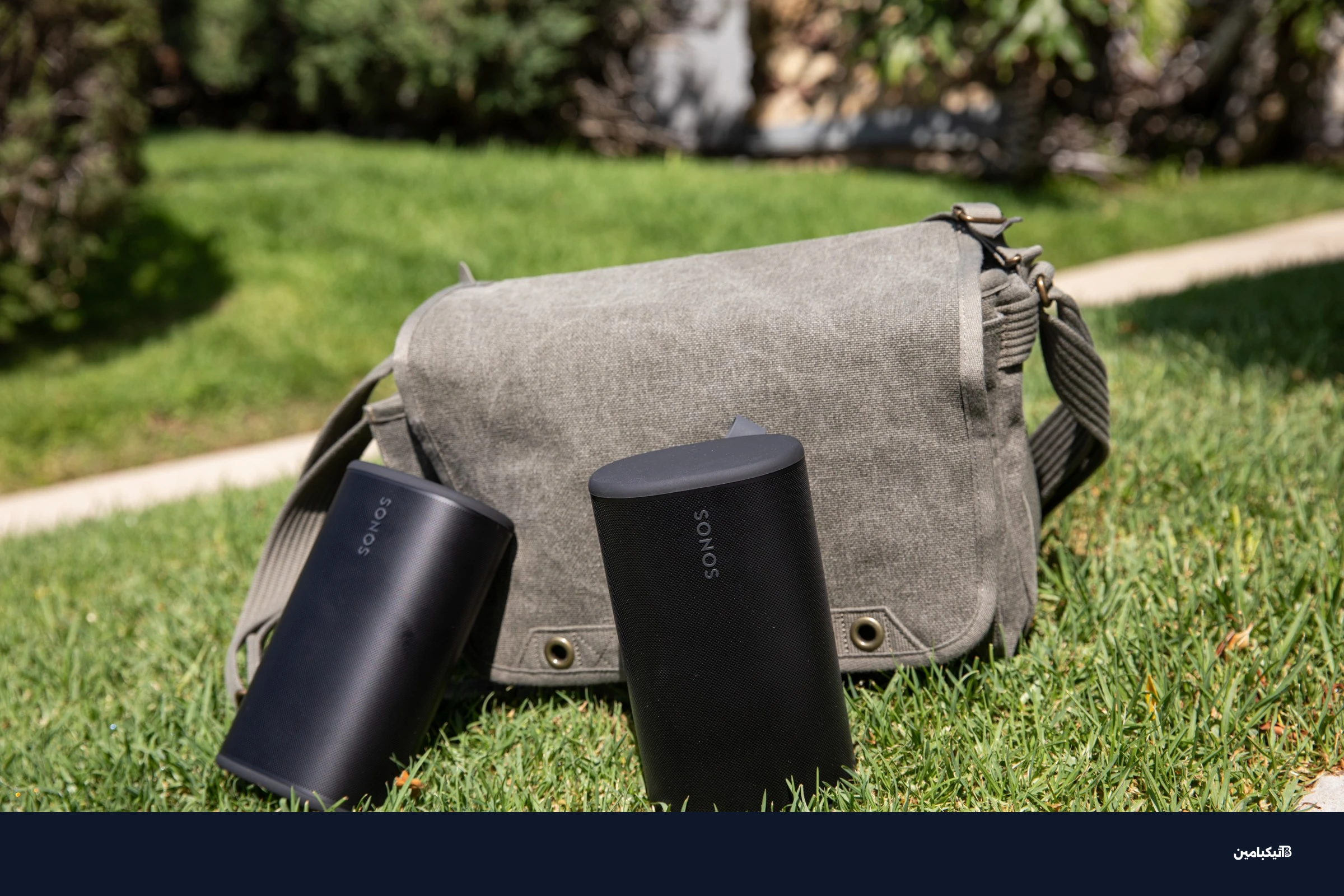 Two Sonos Play speakers in front of a sage colored bag on the grass.