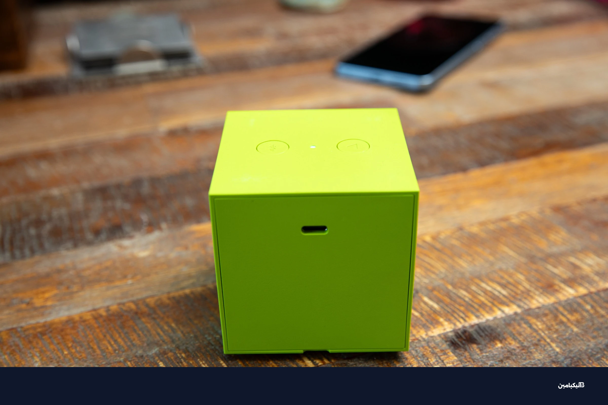 The back of a green Ikea Kallsup speaker on a wooden table, showing its USB-C charge port and two buttons.